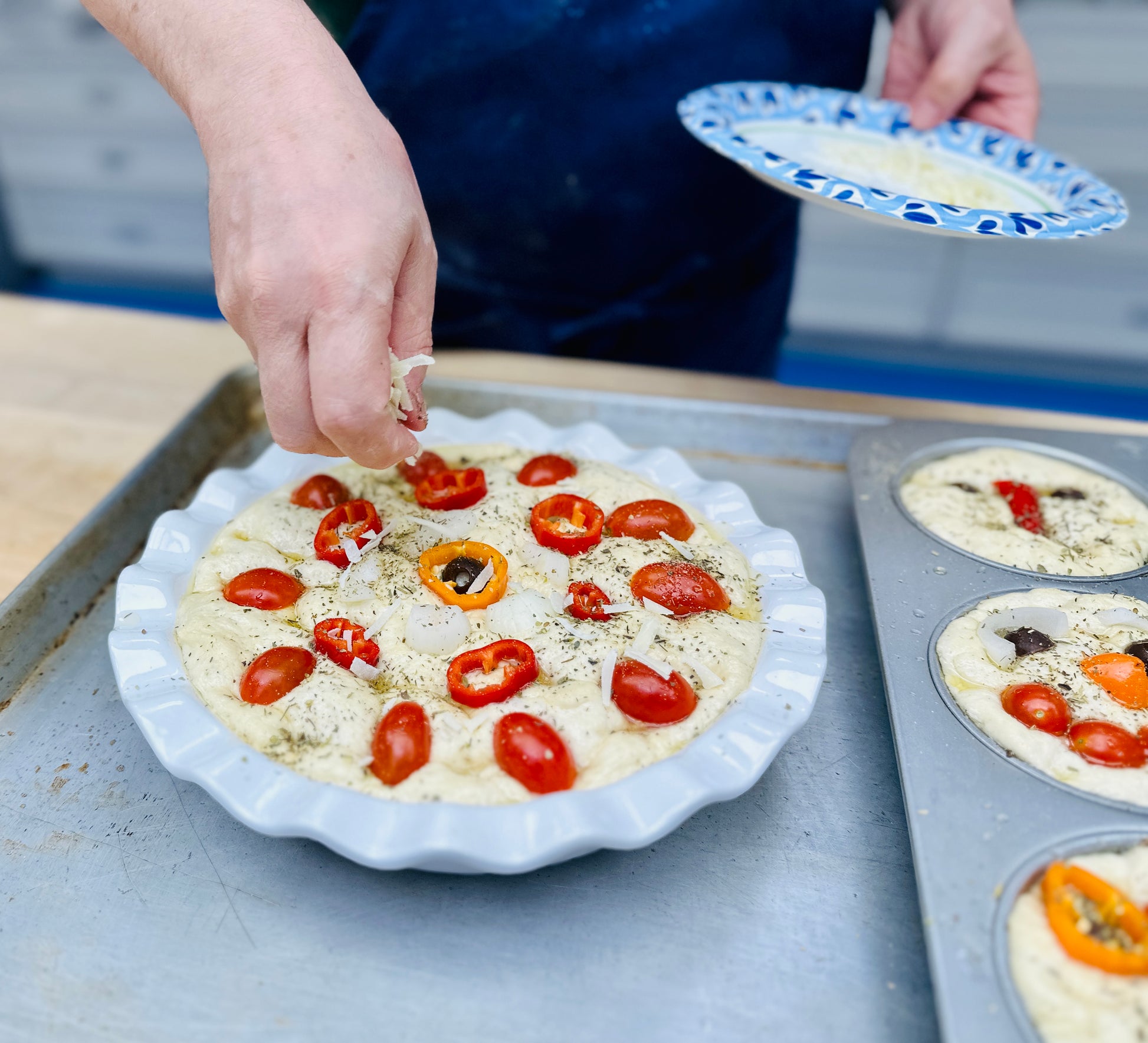A person's hands sprinkle toppings onto focaccia dough