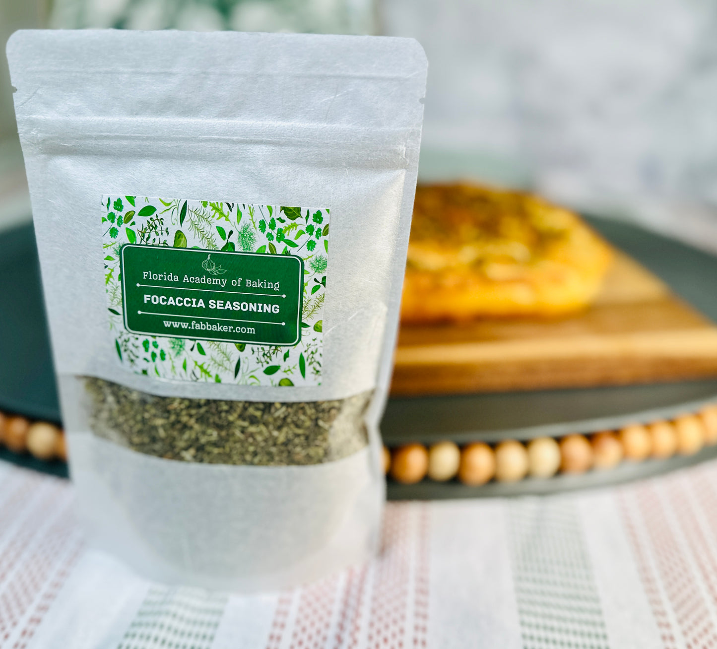 A white resealable pouch of Florida Academy of Baking Focaccia Seasoning sitting next to a freshly baked focaccia bread on a wooden board.
